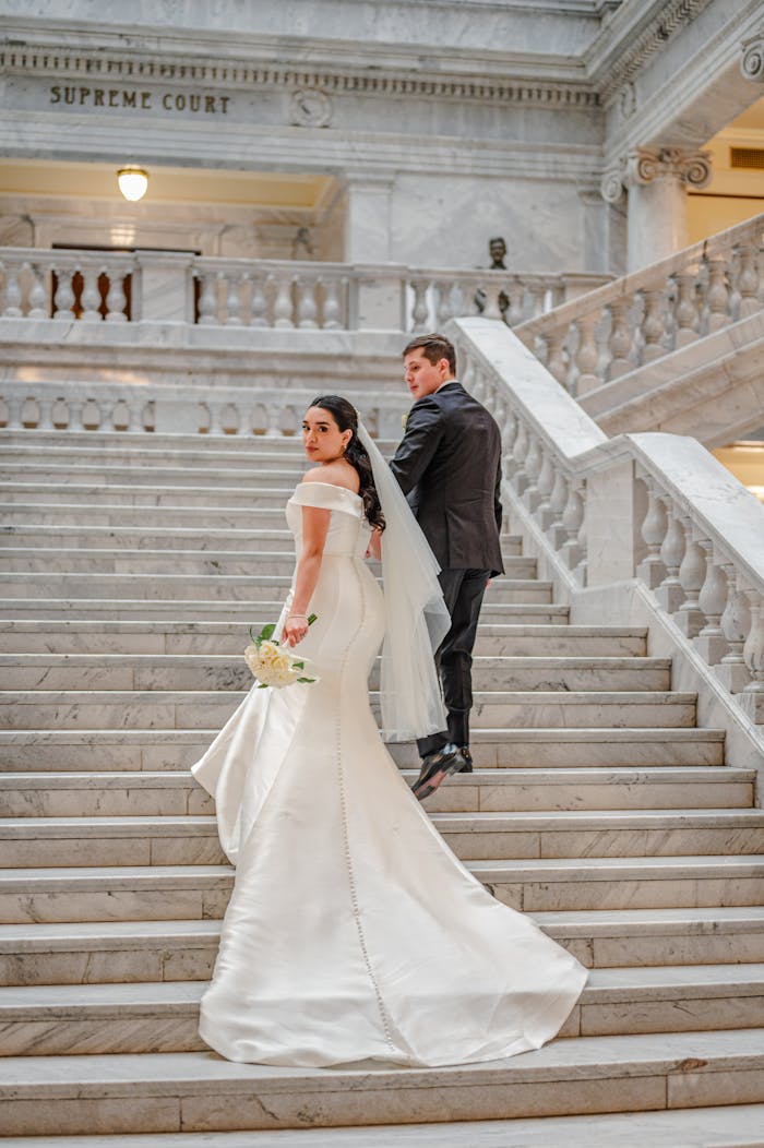 services-03 A bride and groom in formal attire pose on a grand marble staircase inside a historic building.